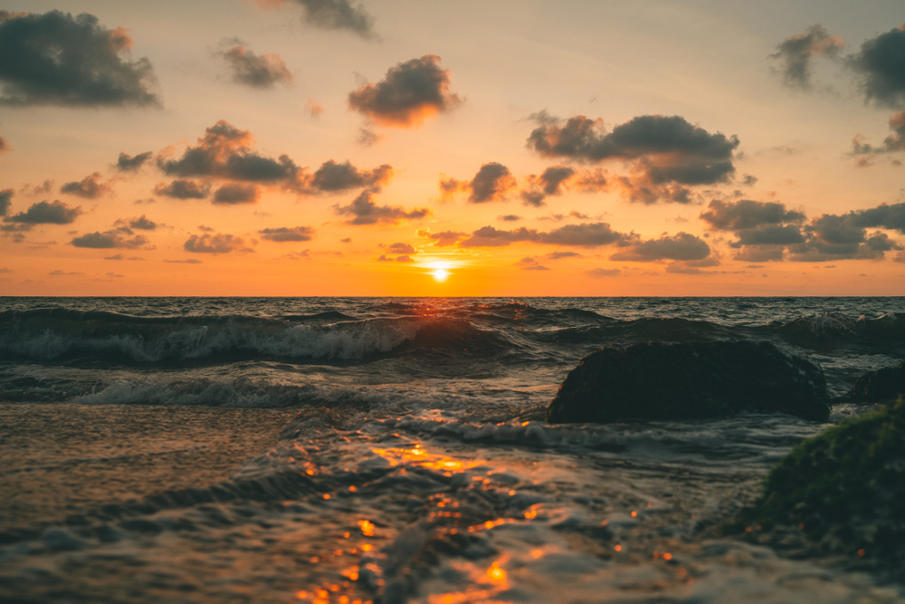 The sun sets beautifully over the ocean, waves crashing on the shore, viewed from Mon Ami City Hotel, evoking tranquility.